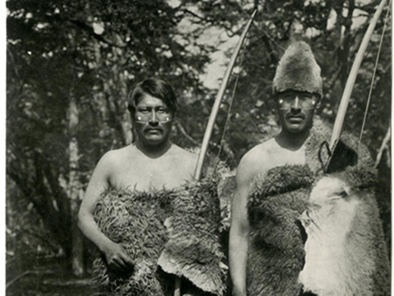 Grupo de mujeres y hombres selk’nam vistiendo pieles de guanaco y portando arcos de caza.